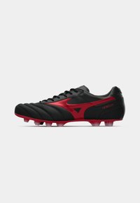 Chuteira de futebol preta com detalhes em vermelho, apresentando um cabedal sintético texturizado, sistema de atacadores, e seis cravos para terreno firme.