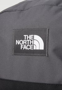 Grå stoffryggsekk med en fremtredende svart og hvit logo-patch. Har en teksturert overflate og kontraststing.