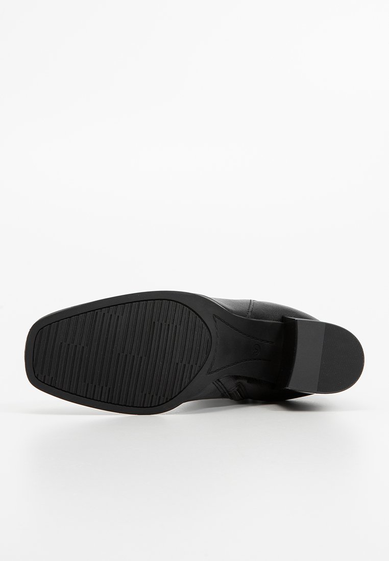 Une chaussure en cuir noir couchée sur le côté, montrant la semelle en caoutchouc à motifs et le talon carré sur un fond blanc.