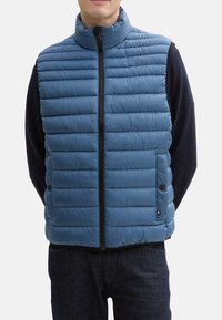 Blauwe gewatteerde vest met verticale gestikte patroon, hoge kraag, volledige rits aan de voorkant en zijzakken. Draagt over een zwart langemouwen top.