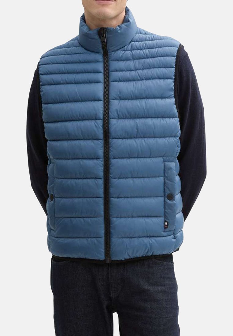 Blauwe gewatteerde vest met verticale gestikte patroon, hoge kraag, volledige rits aan de voorkant en zijzakken. Draagt over een zwart langemouwen top.