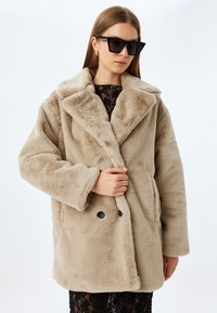 Abrigo de piel sintética beige con un gran cuello, botones de doble botonadura y bolsillos laterales. La modelo lleva gafas de sol negras de gran tamaño y una blusa con detalles de encaje debajo.