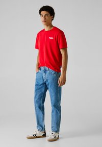 Joven de pie, vistiendo una camiseta roja de Red Bull Racing, jeans de color azul claro y zapatillas blancas con detalles en negro y marrón.