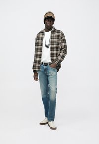 Plaid brown and cream button-up shirt over a white t-shirt, light blue jeans, cream sneakers, and a multicolored beanie. Black accessory around neck.