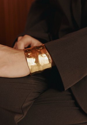 Zlati manšetni bracelet z teksturirano, valovito površino, nosi se na zapestju proti temni tkanini v ozadju. Preprost, širok dizajn poudarja sijaj.