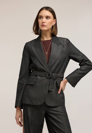 Donna con capelli castani alla lunghezza delle spalle che indossa una giacca elegante grigia scura con cintura e pantaloni abbinati, in posa con una mano sui fianchi.