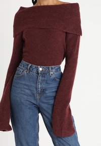 Tmavě červený svetr se spadlými rameny a dlouhými širokými rukávy, žebrovaná textura, v kombinaci s džínami s vysokým pasem.