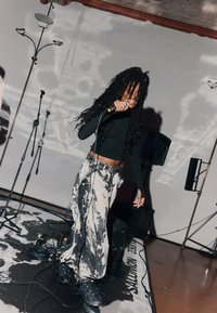 Egy fekete hosszú ujjú felsőt és mintás szürke nadrágot viselő előadó mikrofont tart a kezében, és egy színpadon áll, ahol zenei felszerelések vannak.