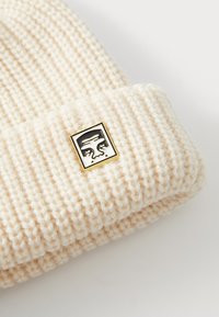 Cream-colored ribbed knit beanie with a folded cuff featuring a small rectangular metal pin showing a stylized black and white face.
