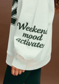 Felpa bianca con testo verde scuro "Weekend mood activated" sulla manica. Presenta un collo rotondo e polsini a costine.