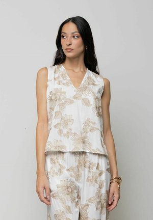 Donna con capelli lunghi e scuri che indossa una canotta bianca senza maniche e pantaloni coordinati con motivi floreali beige, in piedi davanti a uno sfondo chiaro uniforme.