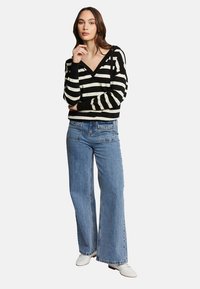 Pull rayé noir et blanc avec un col en V, associé à un jean large bleu clair et des chaussures blanches. Le mannequin se tient les mains sur le menton.