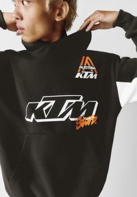 Čierna mikina s kapucňou s bielym a oranžovým logom "KTM", s vysokým golierom a dvoma tónmi rukávov v bielej a čiernej farbe. Detail predného vrecka.