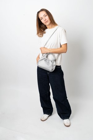 Donna che indossa una maglietta bianca e jeans larghi scuri, tiene in mano una borsa grigia chiara con tracolla, in piedi su uno sfondo bianco.
