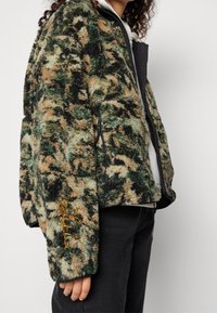 Kurtka polarowa w zielonym, brązowym i czarnym wzorze kamuflażu z czarnym zamkiem błyskawicznym i mankietami. Posiada haftowane detale na rękawie.