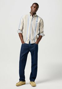 Camisa de botones a rayas en beige claro, azul y blanco; jeans oscuros de corte relajado; mocasines marrones; manos en los bolsillos.
