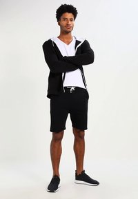 Črna kapa s kapuco z zadrgo in belimi poudarki, bela majica z V-izrezom in črne kratke hlače. Celoten videz dopolnjujejo črni športni copati.