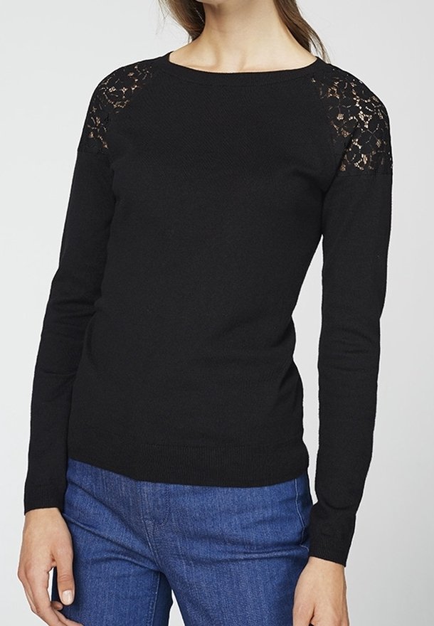 Pull noir à manches longues avec détails en dentelle sur les épaules porté avec un jean bleu par une personne debout sur un fond uni.