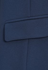 Ύφασμα σε ναυτικό μπλε με υφή πλέξης, που διαθέτει επίπεδη τσέπη με στρογγυλεμένη λεπτομέρεια στο καπάκι. Το υλικό φαίνεται απαλό και δομημένο.