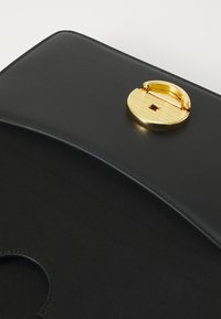 Borsa a spalla in pelle nera con una texture liscia, dotata di una chiusura rotonda in oro con superficie rigata e un dettaglio di ritaglio interno.