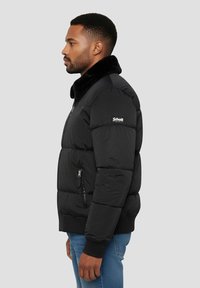 Veste matelassée noire avec col en fausse fourrure, poche latérale zippée, poignets élastiques et ourlet inférieur ajusté. Patch logo sur l'épaule.