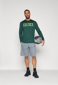 Žalia ilgos rankovės marškiniai su užrašu "CELTICS", pilkos šortai ir juodi sportbačiai. Laikantis krepšinio kamuolys su tekstūruota paviršiumi, stovintis ant baltos fono.