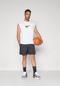 Férfi ujjatlan fehér Nike pólóban, fekete rövidnadrágban és sportcipőben, aki egy narancssárga kosárlabdát tart egy sima szürke háttér előtt.