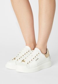 Sneaker bianche con accenti dorati, caratterizzate da una tomaia in pelle liscia, suola spessa, punta rotonda e logo prominente sul lato.