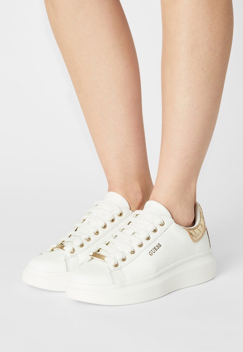 Sneaker bianche con accenti dorati, caratterizzate da una tomaia in pelle liscia, suola spessa, punta rotonda e logo prominente sul lato.