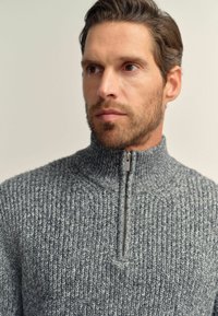 Grauer, mit Reißverschluss versehener Strickpullover mit hohem Kragen. Der Stoff weist ein strukturiertes Rippenmuster und einen subtilen Glanz auf.