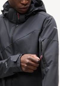 Άτομο που φοράει μαύρο μπουφάν με κουκούλα ανοίγοντας μια τσέπη με φερμουάρ στο πάνω μανίκι, δείχνοντας λεπτομέρειες και υφές του μπουφάν.