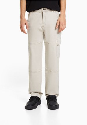 Beige straight-leg cargo pants with knee seams and flap pocket on right leg, worn with black sneakers and black shirt.