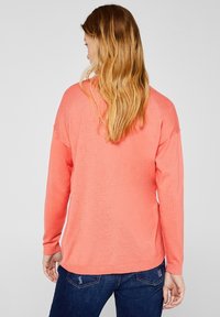 Koraalkleurige pullover met lange mouwen, zachte textuur, verlaagde schouders en een rechte zoom. Gemaakt in combinatie met donkerblauwe jeans.