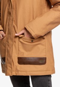 Parka marrón con un acabado texturizado en tono beige, que presenta dos bolsillos con acentos de cuero marrón oscuro y un detalle de botones a presión.
