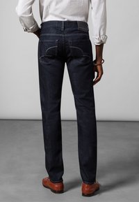 Mörkblå denimjeans med raka ben, som visar kontrasterande sömnad på bakfickorna, bärs med bruna skor och en ljusfärgad skjorta.