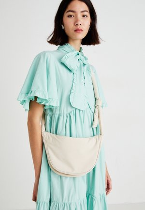 Kvinde med skulderlangt mørkt hår iført en mintgrøn kjole med flæser og bærende en beige skuldertaske mod en ensfarvet hvid baggrund.