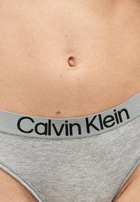 Κοντινή λήψη του κάτω μέρους της κοιλιάς ενός ατόμου που φοράει γκρι εσώρουχο Calvin Klein, δείχνοντας το λογότυπο στη μέση και έναν μικρό σπίλο πάνω από τον αφαλό.