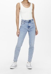 Persona usando una camiseta sin mangas blanca de canalé, jeans de cintura alta azul claro, zapatillas blancas y un collar de cadena de plata sobre un fondo blanco.