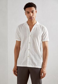 Short-sleeve white shirt featuring eyelet fabric with circular cutouts, a notched collar, and a button-down front. Worn with gray pants.