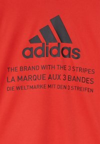 Logo Adidas s tagline "Značka se třemi pruhy" v angličtině, francouzštině a němčině na červeném textilním pozadí.