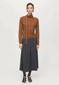Donna che indossa un maglione lungo marrone con cravatta al collo, una gonna midi plissettata grigia scura, collant neri e tacchi bianchi con cinturini, in piedi contro uno sfondo bianco.