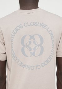 Μπεζ βαμβακερή μπλούζα t-shirt με κυκλική γραφιστική που περιλαμβάνει το κείμενο "CLOSURE STUDIOS LONDON" και ένα στυλιζαρισμένο λογότυπο σε ανοιχτό γκρι στην πλάτη.
