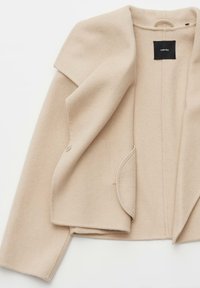 Cappotto beige in misto lana con apertura frontale, ampi risvolti e una tasca singola. L'interno presenta un'etichetta con il nome del marchio.