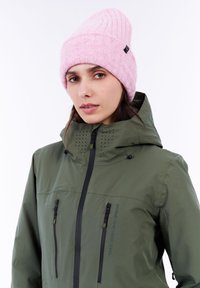 Grüne wasserdichte Jacke mit Reißverschlusstaschen und Belüftungsöffnungen, kombiniert mit einer rosa gerippten Mütze. Glatte Textur, modernes Design.