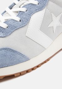 Heled sinine ja hall tennisepoos, mis on valmistatud nahast ja sametist, esitades valge tähise logo, tekstuuriga aksessuaare ja lamedad valged paelad.