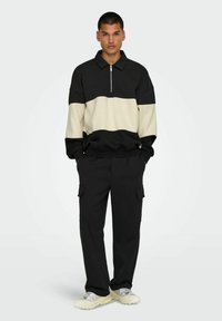 Schwarz- und cremefarbener color-blockierter Halb-Reißverschluss-Pullover, kombiniert mit schwarzen Cargo-Hosen. Der Stoff wirkt weich; der Pullover hat einen Kragen und lange Ärmel.