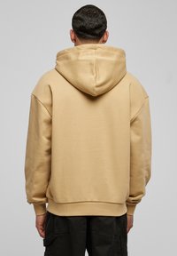 Felpa oversize beige realizzata in tessuto morbido, caratterizzata da un ampio cappuccio e polsini a costine. Texture liscia con cuciture pulite e senza grafiche visibili.