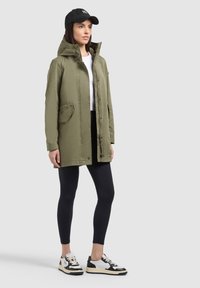 Donna che indossa un parka con cappuccio verde oliva, camicia bianca, leggings neri, cappellino da baseball nero e sneakers bianche e nere, in piedi contro uno sfondo grigio.