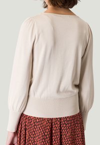 Jasno beżowy sweter z dzianiny o fakturowanej powierzchni, z bufiastymi długimi rękawami i ściągaczem u dołu. Noszony na tle wzorzystej czerwonej spódnicy z okrągłymi motywami.