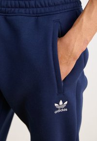 Tumši zilas sporta bikses ar maigu tekstūru, kas aprīkotas ar sānu kabatām un baltu Adidas logo kreisajā augšstilbā. Iekļauta elastīga josta.